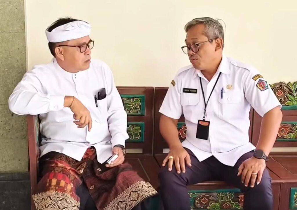 Whatsapp image 20260607 at 21 17 42 catrawarta Two men sit on a wooden bench indoors mid conversation the man on the left wears a white traditional outfit and headband with a patterned sarong and sandals the man on the right wears a white uniform shirt with a name badge and lanyard
